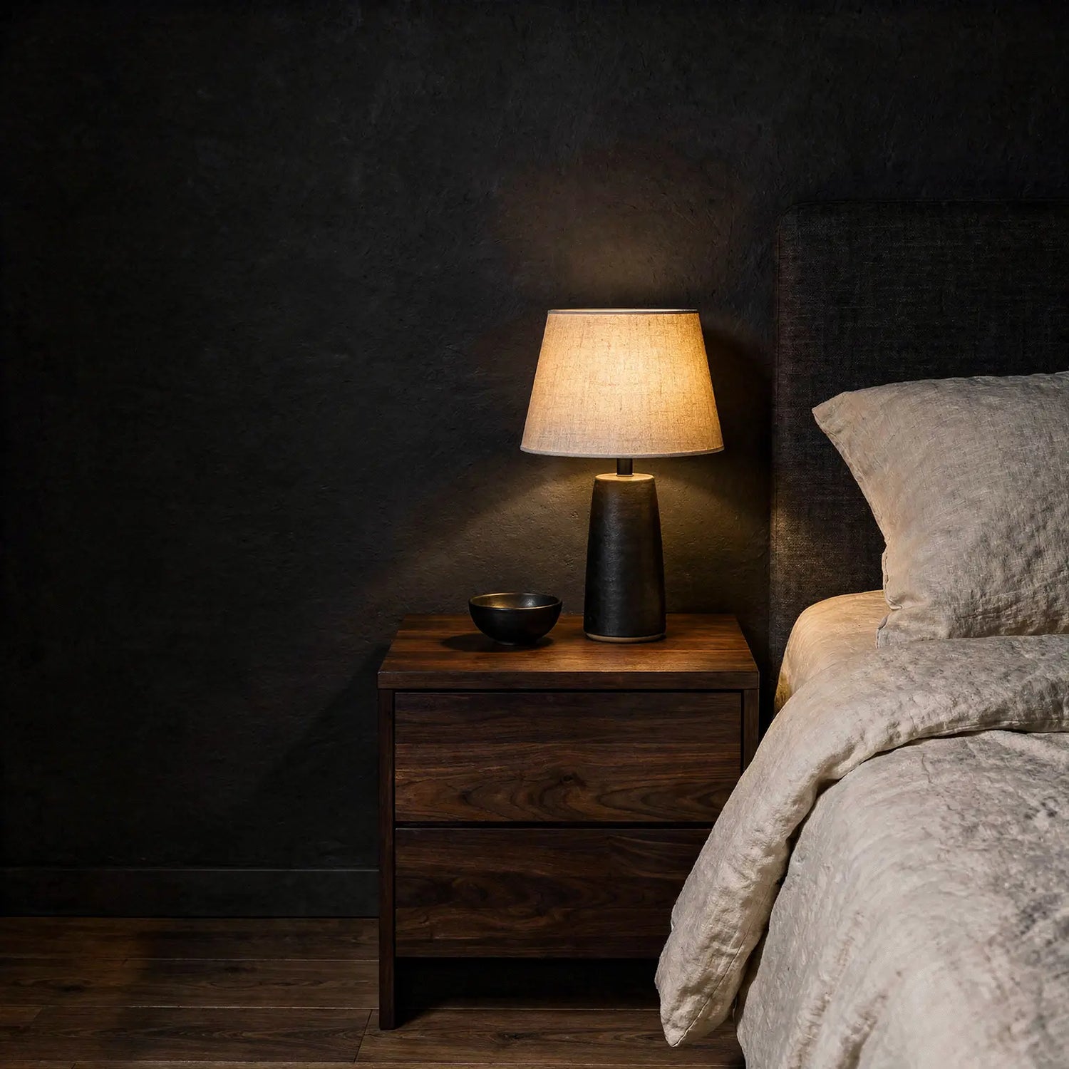 Warm bedside lamp on a dark walnut nightstand beside ivory bedding