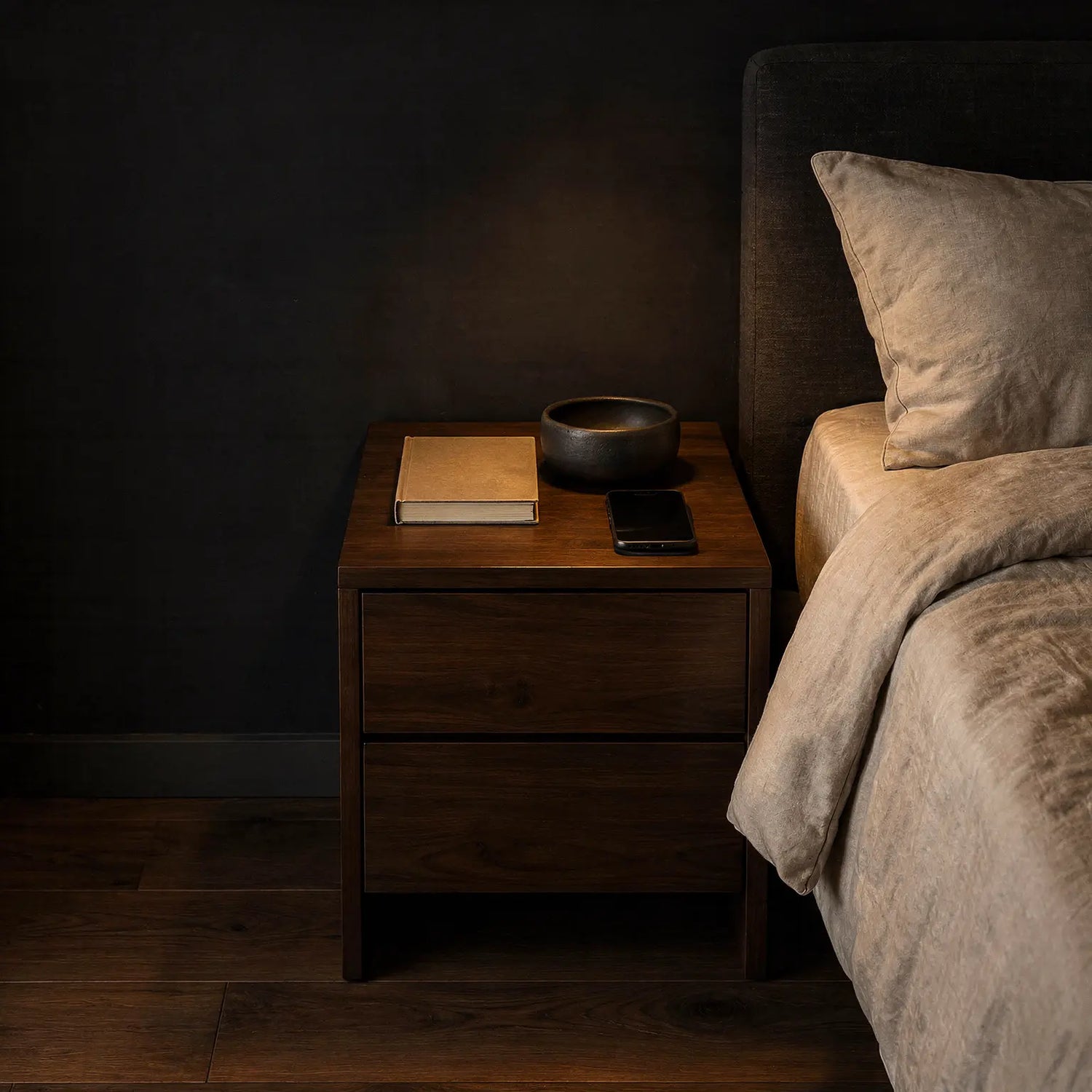 Dark walnut nightstand with a closed neutral book, matte bowl, and black smartphone beside ivory bedding