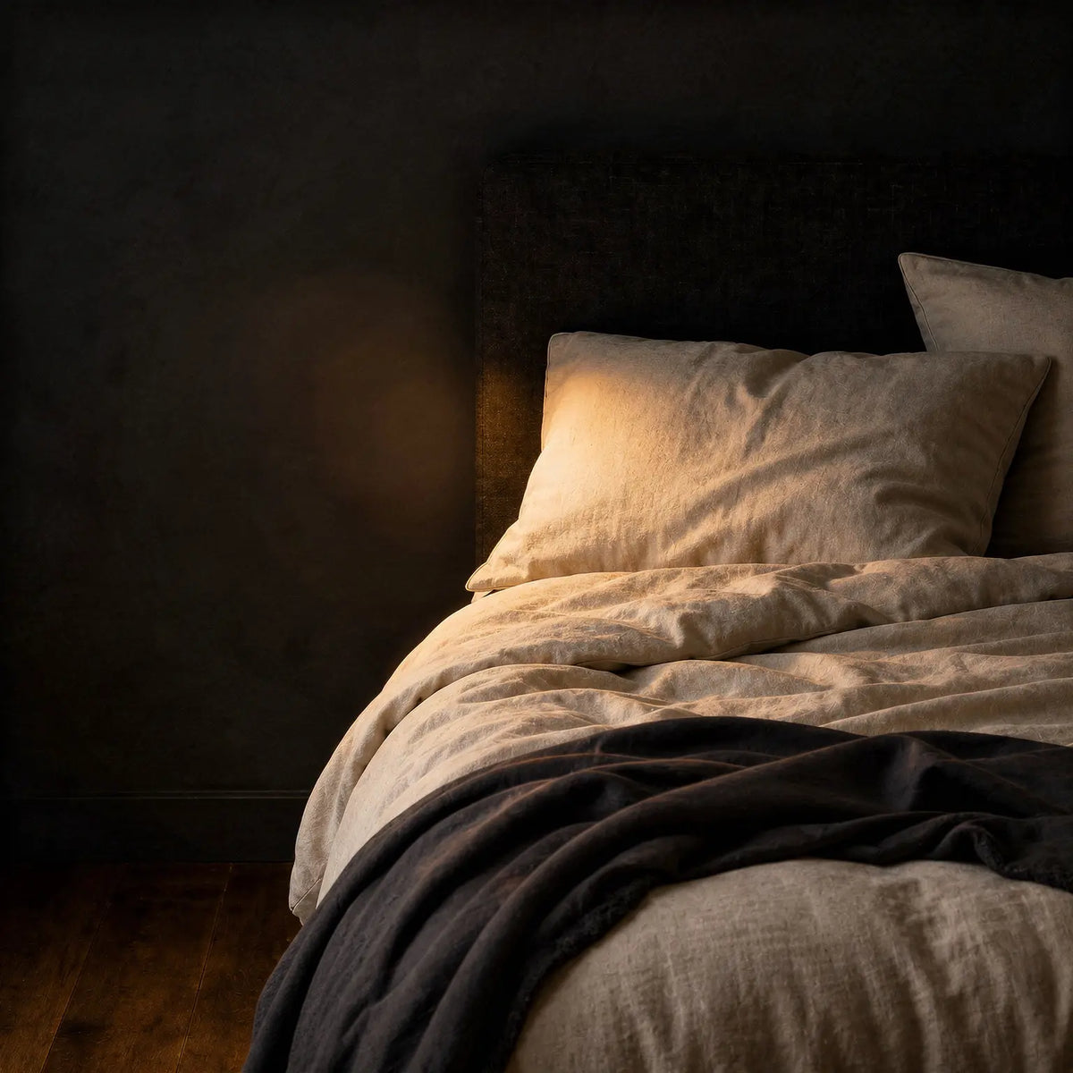 Warm ivory linen bedding with a dark charcoal throw against a dark upholstered headboard